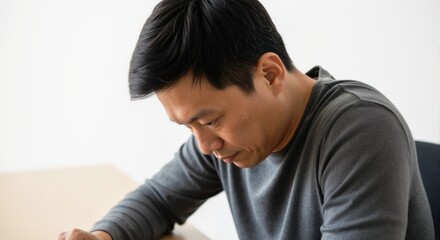 Asian man sitting with a serious expression while looking down at table  