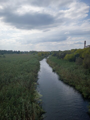 Fototapeta premium Serene river flows through lush green landscape under a cloudy sky, surrounded by vibrant foliage and distant structures, showcasing natural beauty and tranquility
