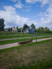 Scenic view of a modern house surrounded by greenery, with a pathway and benches, under a blue sky with fluffy clouds, creating a tranquil outdoor atmosphere