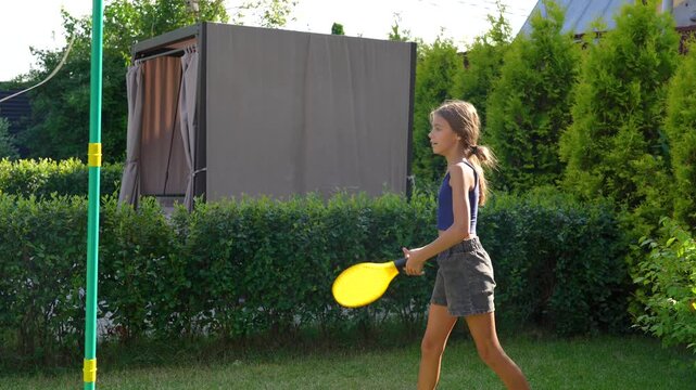 Young girl's energetic tetherball game with paddle in vibrant, sunny summer garden, full of joyful activity.