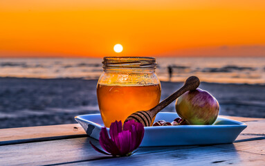 Close up with jar of honey and apples  - traditional food for Jewish New Year holiday - Rosh Hashanah. Focus on food jar with honey, blurred sand beach with people silhouette, sea waves and sunset