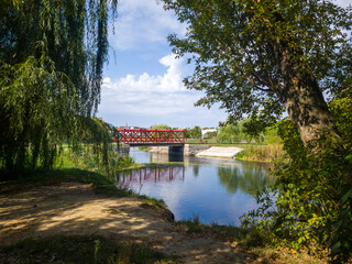 Scenic view of a tranquil river surrounded by lush greenery, featuring a vibrant red bridge, reflecting in the water, creating a serene natural landscape