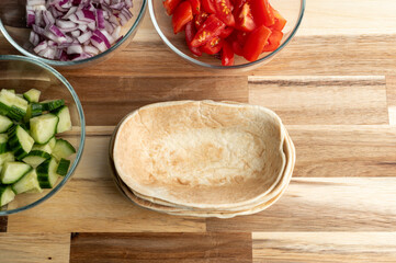 A wooden table with bowls of chopped vegetables including cucumbers, tomatoes, and red onions, alongside empty tortilla shells