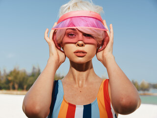 Young woman wearing a rainbow striped swimsuit and pink visor stands on a sunny beach, delivering...