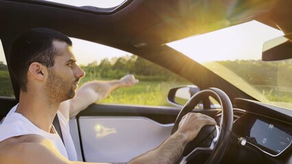 Profile of young handsome man driving a modern electric car with sunlight at background. Young male driver riding on electrical vehicle at countryside. Concept of journey and trip. Close up
