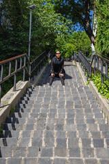 Fototapeta premium A fashionable woman in black outfit and sunglasses poses on a long stone staircase surrounded by lush greenery on a sunny day, radiating strength and confidence.