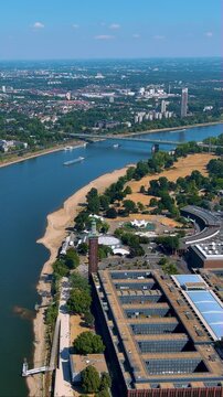 4K vertical drone video sweeps Cologne&rsquo;s Rhine waterfront: Koelnmesse halls, Deutz beach park, bridge and passing ships under clear summer sky - vibrant footage for travel and trade - fair stories.