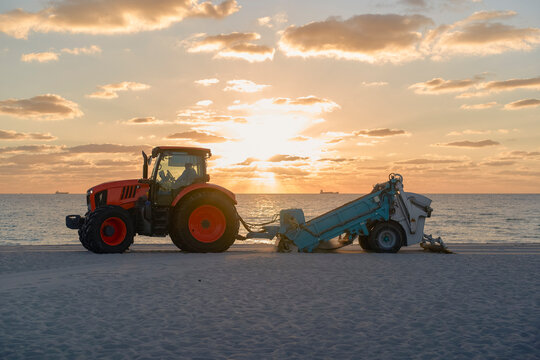 A tractor pulls a sand cleaner on a beach at sunset with the ocean in the background. Miami, Florida, USA