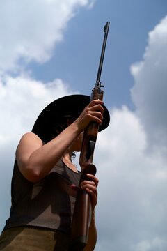 Woman in cowboy hat holds a rifle against a cloudy sky background.
