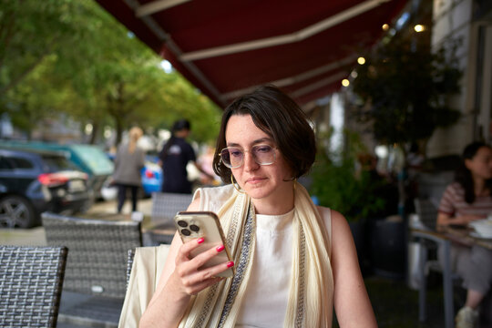 Woman in glasses using smartphone at an outdoor cafe with red awning. Berlin, Germany