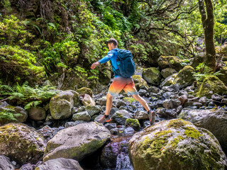 Obraz premium A sporty hiker jumps over river with big boulders in the rainforest. The rainforest is located in São Vicente on the Portuguese island of Madeira.