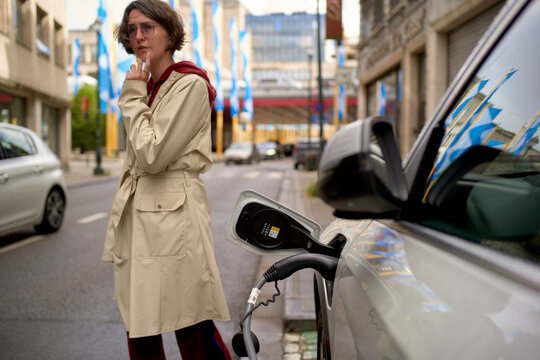 Woman in a trench coat stands by an electric car charging on a city street. Brussels, Belgium
