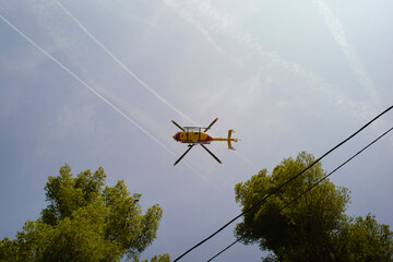 Helicopter flying overhead with trees and power lines in the foreground against a blue sky. Marseille, France