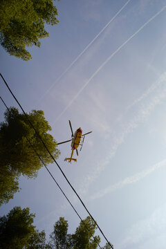 A yellow helicopter flies above trees with contrails in a clear blue sky. Marseille, France