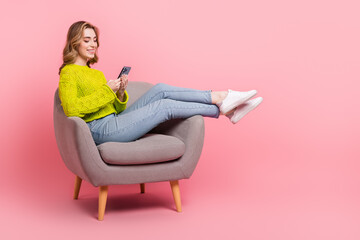 Lovely young woman in casual attire seated on chair using smartphone on bright pink backdrop radiates cheerful energy