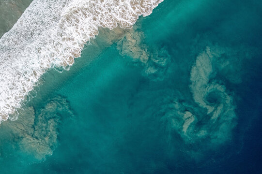 Aerial view of ocean waves crashing on sandy beach with swirling water patterns. South Nias, North Sumatra, Indonesia