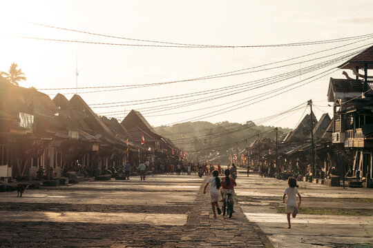 Children playing on a rustic street lined with traditional wooden houses at sunset. Desa Hilisimaetano in South Nias, North Sumatra, Indonesia