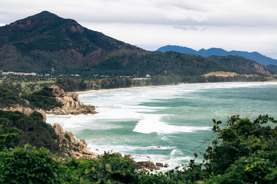 Lush green hills meet a rugged coastline under a cloudy sky with waves crashing on the shore. Ninh Hai District in Ninh Thuan, Vietnam