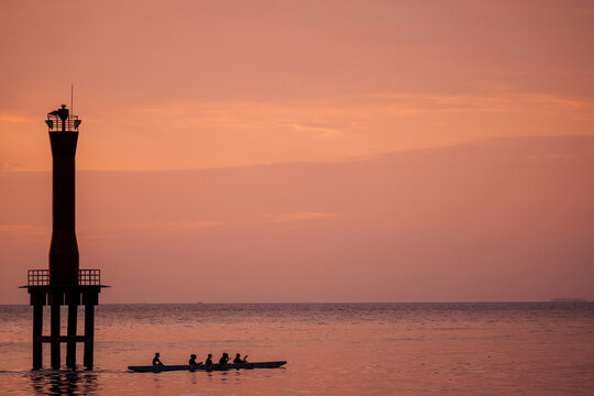 Silhouetted people row a canoe on calm water near a lighthouse at sunset. Padang, West Sumatra, Indonesia 