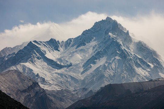Snow-covered mountains with jagged peaks rise majestically under a cloudy blue sky. Road F042, Annapurna, Mustang District, Gandaki Province, Nepal
