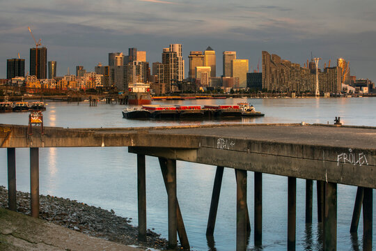 Urban skyline at sunset reflected on a calm river with a concrete bridge in the foreground. Canary Wharf, London, United Kingdom