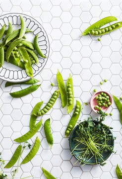 Fresh green peas and pea shoots on a white hexagonal tile surface with bowls.