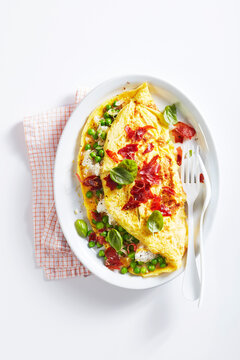 Fluffy omelette with cheese, ham, and green peas garnished with fresh basil on a white plate.