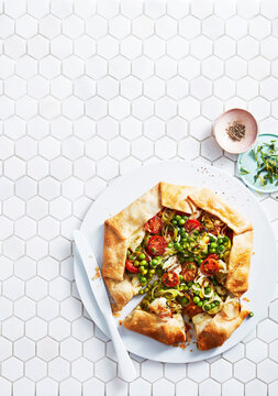 Rustic vegetable galette with tomatoes and peas on white hexagonal tile background.