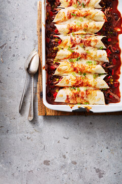 A baking dish with cheesy enchiladas topped with green onions and a side of spoons on a table.