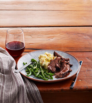 Plate of roast beef, mashed potatoes, green beans, and red wine on wooden table with napkin.