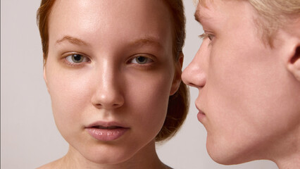 Fototapeta premium Red-haired woman facing camera while blond man leans close in neutral studio portrait. Concept of individuality, intimacy, emotional balance, modern editorial beauty, and connection.