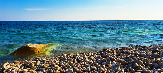 Scenic view of a rocky seashore with clear turquoise water, sunlit waves, and submerged stones creating a vibrant coastal landscape under a bright sky.