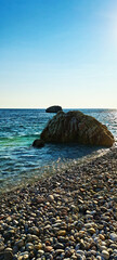 Scenic view of a rocky seashore with clear turquoise water, sunlit waves, and submerged stones creating a vibrant coastal landscape under a bright sky.
