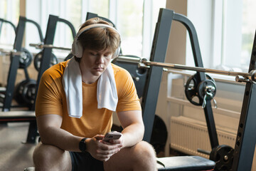 Sporty young man relax and sitting with mobile phone, listen music headphones and texting at gym. Fitness lifestyle concept. Take a break.