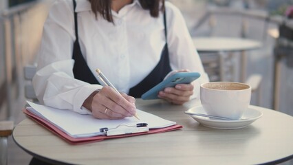 A professional woman sits at a cafe, writing notes on a clipboard while checking her phone. She enjoys a cup of coffee in a relaxed outdoor setting, focused on her tasks