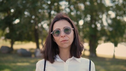 A confident businesswoman stands in a green park wearing sunglasses. She gazes forward with a thoughtful expression. The sunlight filters through the trees, enhancing her professional look