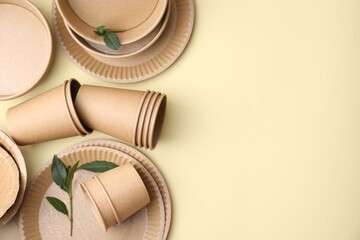 Set of disposable tableware and green leaves on beige background, flat lay. Space for text