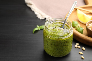 Fresh pesto sauce with spoon in jar and ingredients on black wooden table, closeup. Space for text