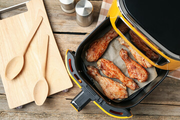 Air fryer with cooked chicken drumsticks and kitchen utensils on wooden table, flat lay