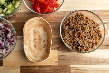 A wooden table with various ingredients for cooking, including chopped vegetables like cucumber and tomatoes in bowls, and a bowl of cooked ground meat, with an empty dish nearby