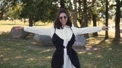 A woman stands in a park with arms wide open, smiling brightly. She wears stylish sunglasses and a trendy outfit. The greenery and rocks in the background add to the cheerful atmosphere