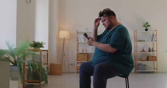 Fat overweight man with glasses holds smartphone, squinting at screen as he tries reading, struggling for focus and better vision. Man with farsightedness eye disease squints at phone screen