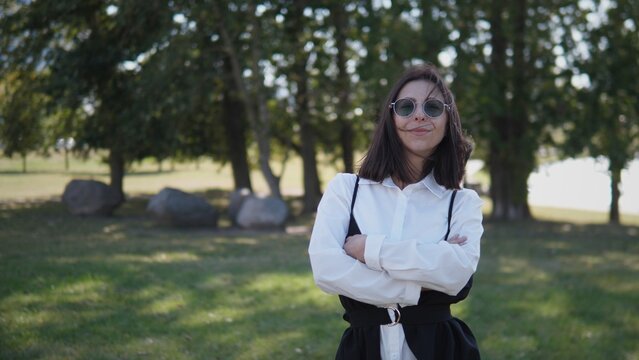 A confident businesswoman poses with arms crossed in a park, wearing sunglasses and a stylish outfit. Trees and rocks provide a natural backdrop in the bright sunlight - Powered by Adobe