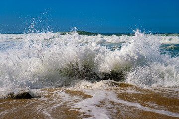 Wave breaking on a granite rock on Matosinhos beach in Portugal. The force of the impact creates drops, splashes, and foam. Shot of the surf at high tide from a low angle. Sandy beach near Porto city.