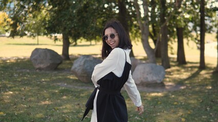 A businesswoman stands outdoors in a park, wearing stylish sunglasses and a casual outfit. She smiles brightly while surrounded by greenery and large rocks, enjoying a sunny day