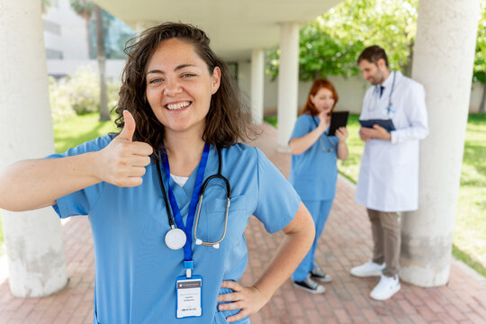 Healthcare worker smiling, giving thumbs up, showing approval and success with medical team members in background