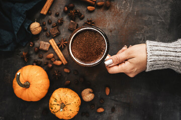 Top view of female hand holding mug of hot chocolate, pumpkins and spices on rustic background, cozy autumn scene