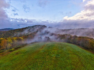 Drone aerial view of fog dissolving over a hill with trees and foliage in the morning with massiv cloud formations. 