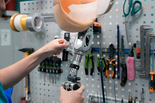 A technician works on a prosthetic leg using a screwdriver. The limb is placed on a workbench with various tools hanging in the background