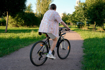 Fototapeta premium A person rides a bike down a paved path. Trees line the sides of the path in a grassy area of the park. It is a sunny day in the park, ideal for a relaxing ride